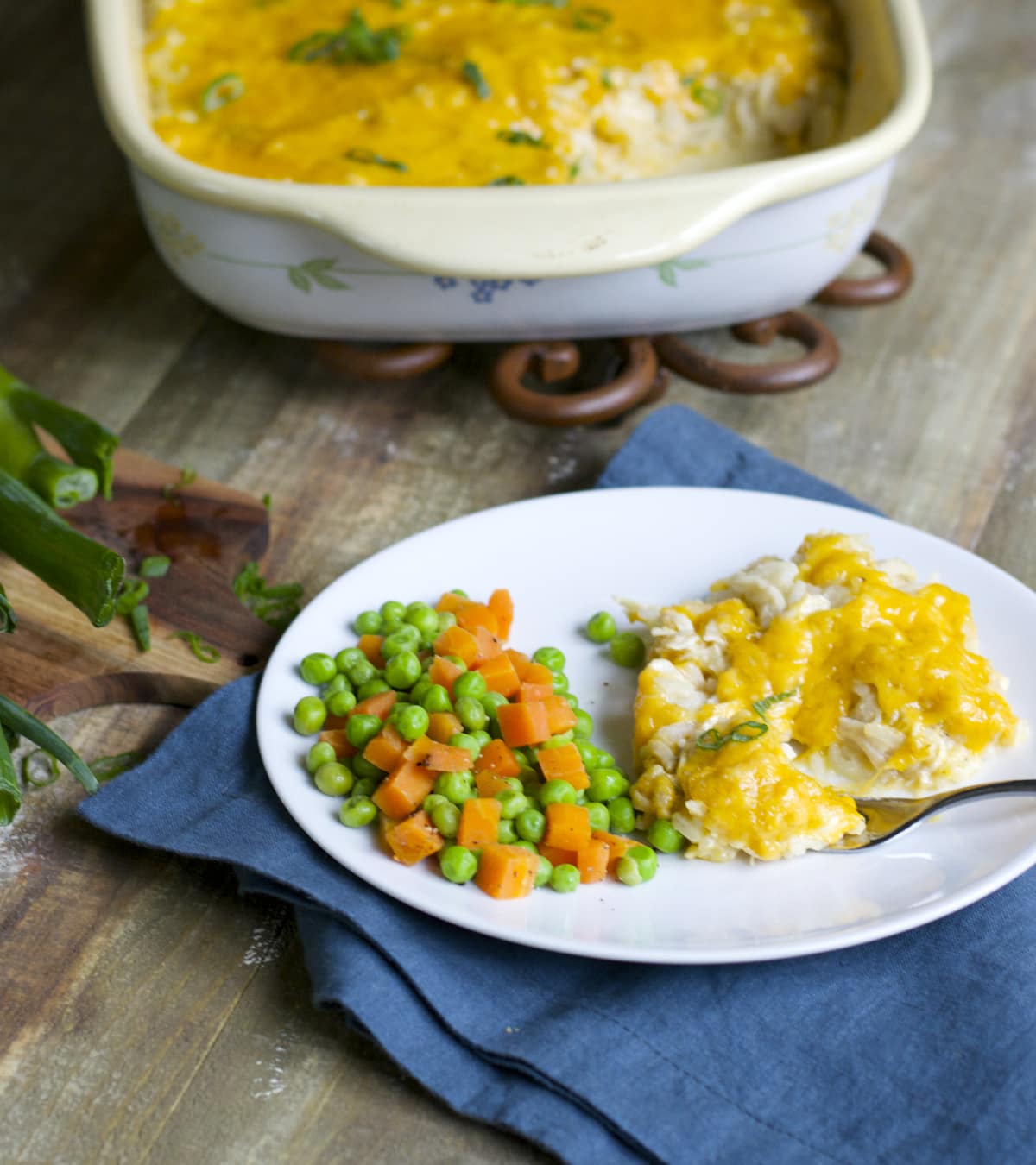 serving of cheesy chicken and rice casserole on a white plate with mixed peas and carrots. A baking dish of casserole rests in the background. 