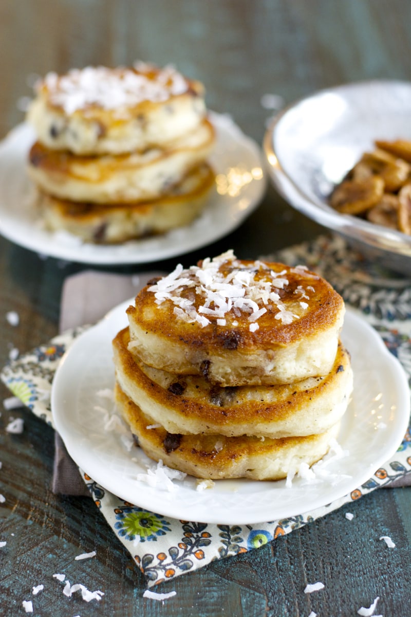 three homemade chocolate chip pancakes on a white plate. Another stack of pancakes is in the background. 