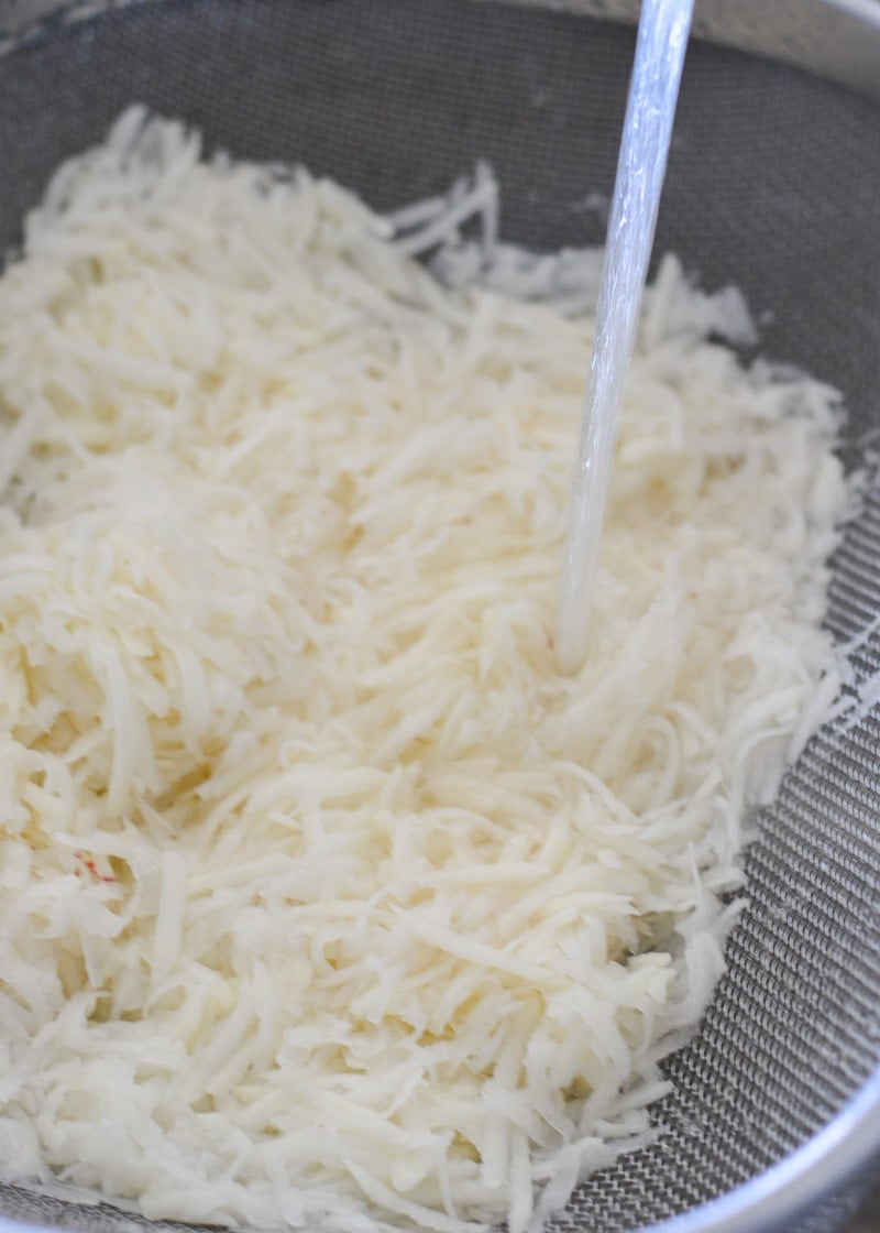 Shredded potatoes in a strainer being run under tap water.