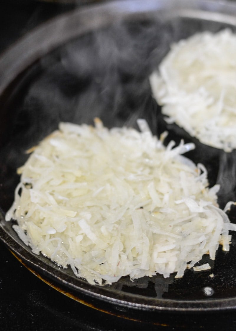 Two homemade hash browns in a hot skillet, being cooked. 