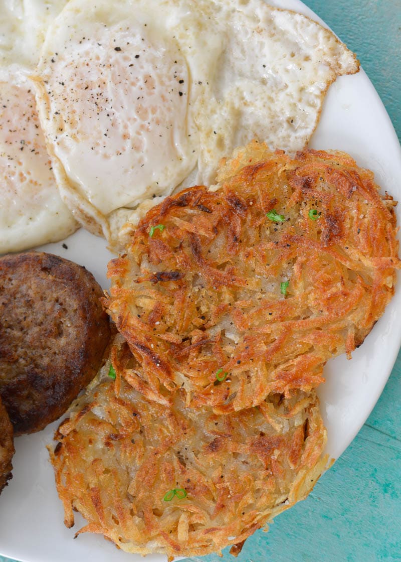 Two homemade hash browns on a white plate with a sausage and two fried eggs. 