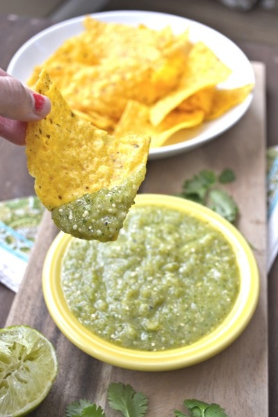 A chip laden with roasted tomatillo salsa, being held over a dish of salsa. 