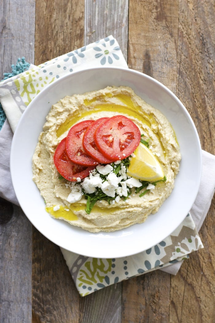 overhead shot showing a bowl of creamy feta hummus topped with a drizzle of oil, tomato slices, fresh herbs, feta, and lemon