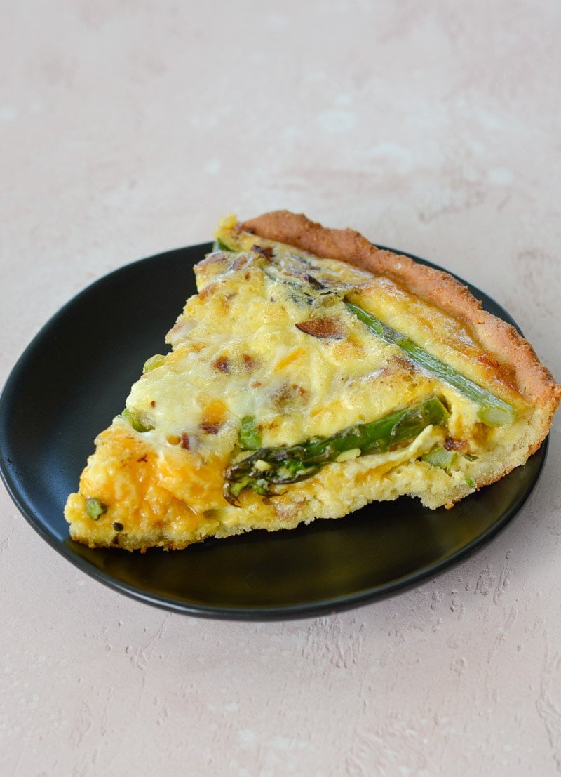 A slice of asparagus, bacon and cheddar quiche on a gray dessert plate. 