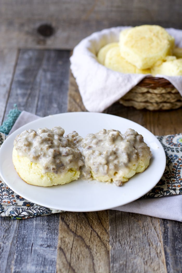 a biscuit split in half and topped with homemade sausage gravy on a white plate. A basket of biscuits rests in the background. 
