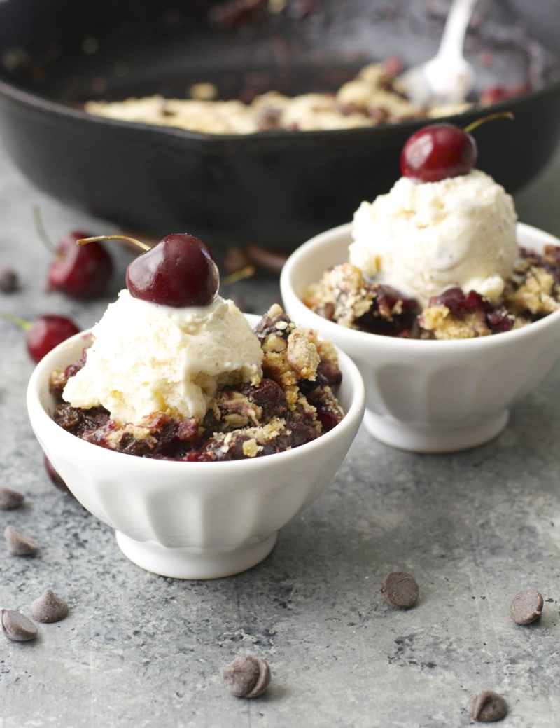 Two white bowls with cherry crisp. A cast iron skillet in the background. 