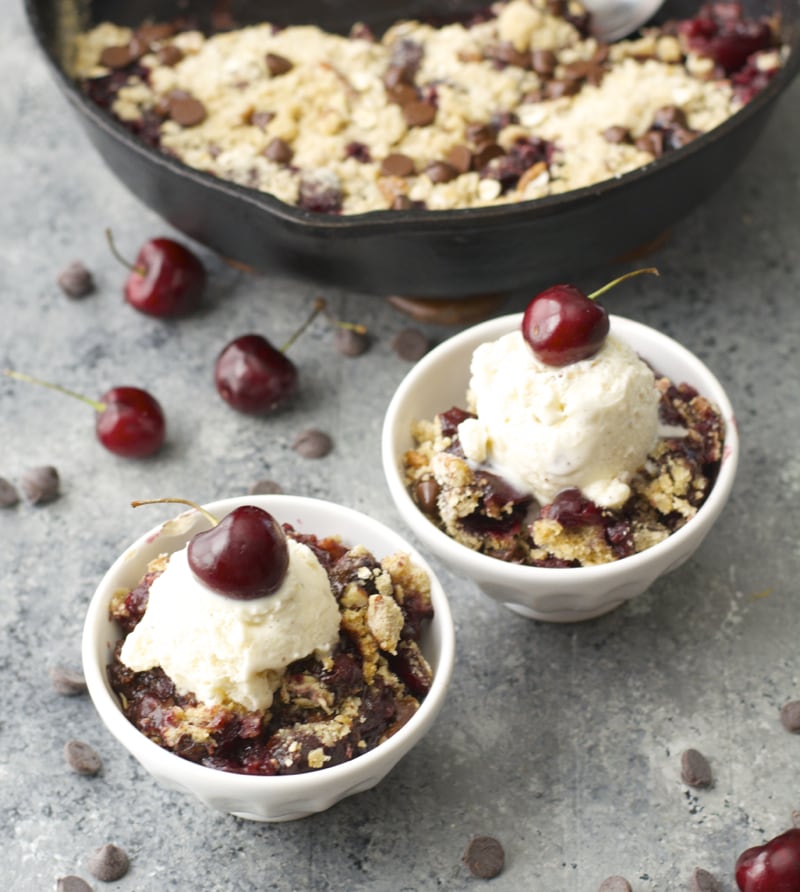 two bowls of cherry crisp with ice cream on top. A skillet of fruit crisp rests in the background. 