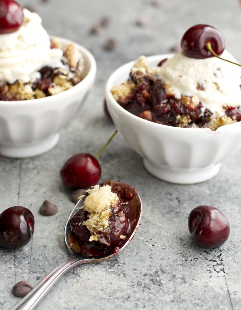 Two bowls of fresh cherry crisp a la mode with a spoon resting in the foreground. 