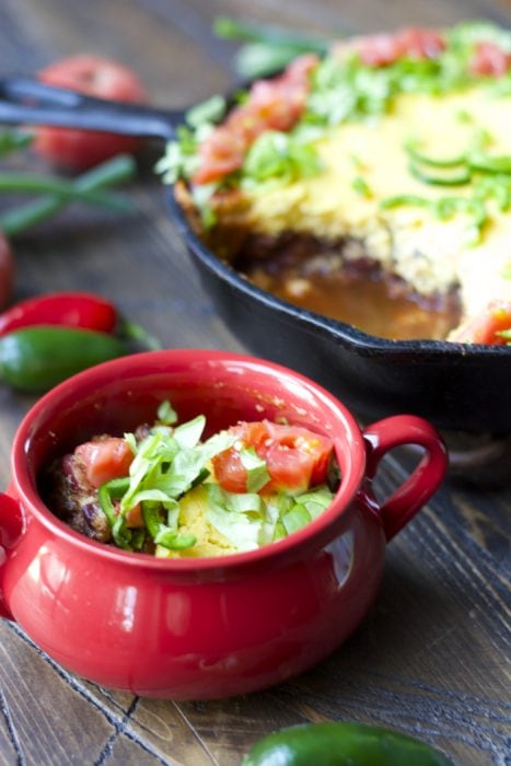 a serving of chili with cornbread topping in a red dish. The entire chili pie rests in the background. 