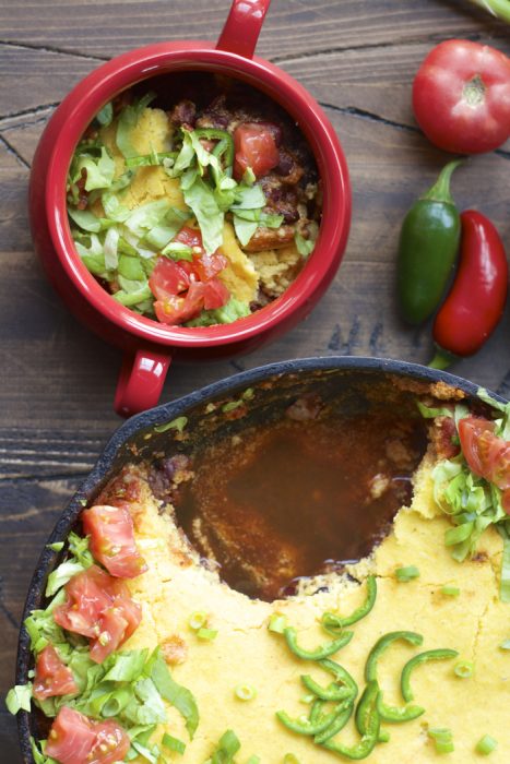 overhead view of chili cornbread casserole in a cast iron skillet. a single serving is in a red bowl next to it. 