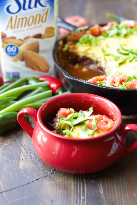a serving of chili cornbread bake in a red bowl. the entire chili pie and a carton of almond milk rest in the background.
