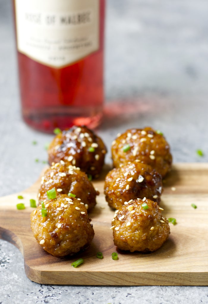 a cheese board with six Asian style meatballs. a bottle of rose wine is in the background. 