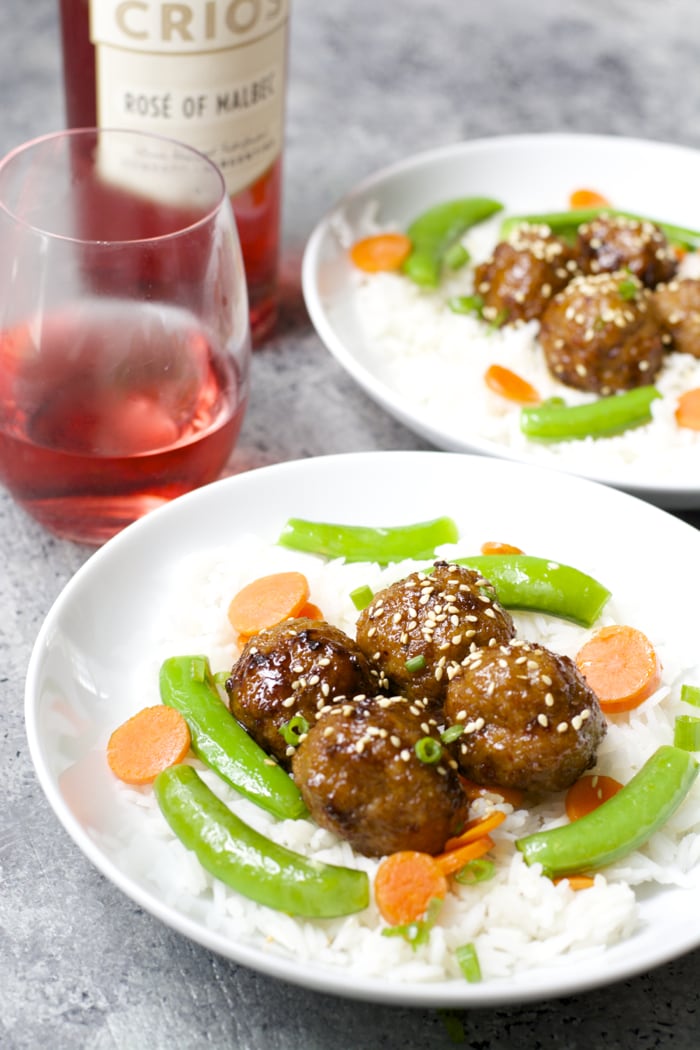 two plates of rice, veggies, and spicy asian meatballs next to a bottle of rose wine 