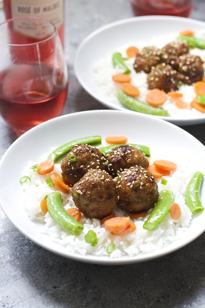 two plates of rice, veggies, and spicy asian meatballs next to a bottle of rose wine 