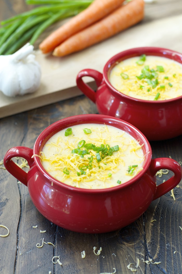 two red bowls filled with homemade potato soup. carrots, green onion, and garlic sit nearby. 