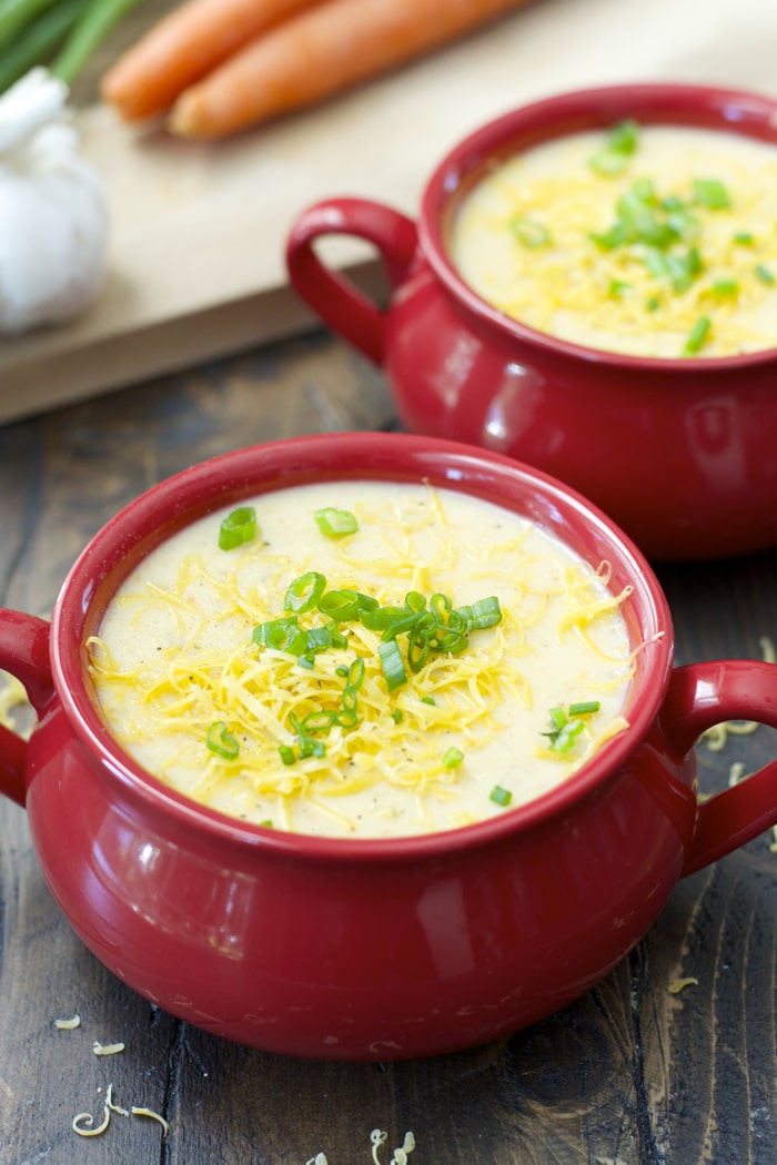 two red bowls filled with crockpot potato soup. carrots, green onion, and garlic sit nearby. 