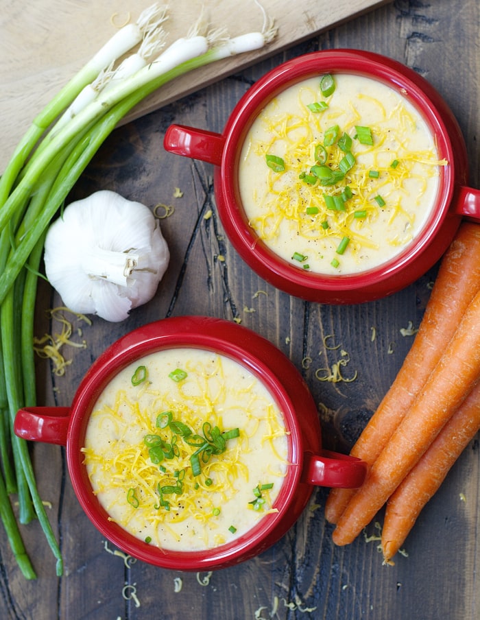 overhead view of two red bowls filled with slow cooker potato soup. carrots, green onion, and garlic sit nearby. 