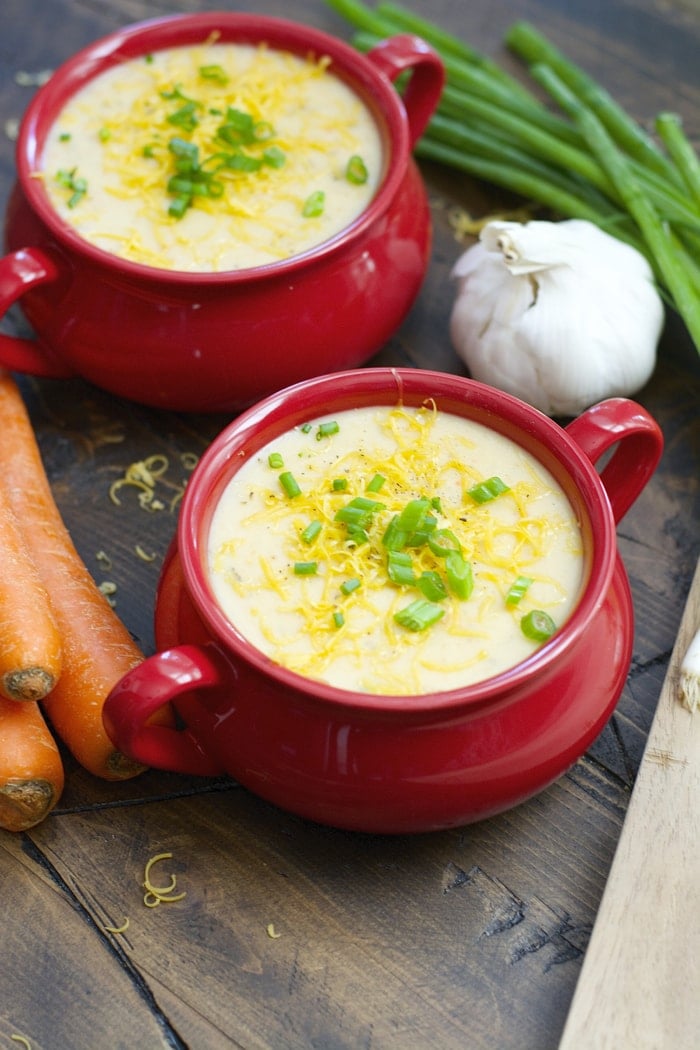 two red bowls filled with slow cooker potato soup. carrots, green onion, and garlic sit nearby. 