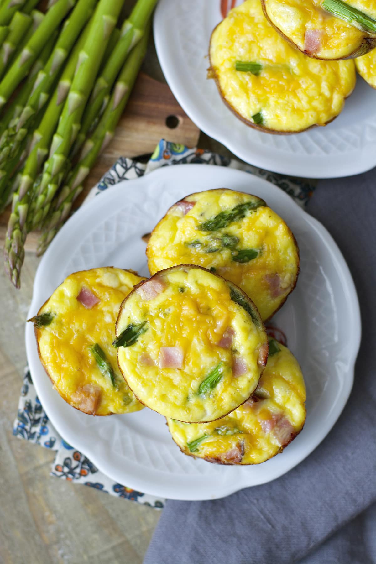 Overhead view of two plates piled with mini ham asparagus quiche, next to a bunch of fresh asparagus. 