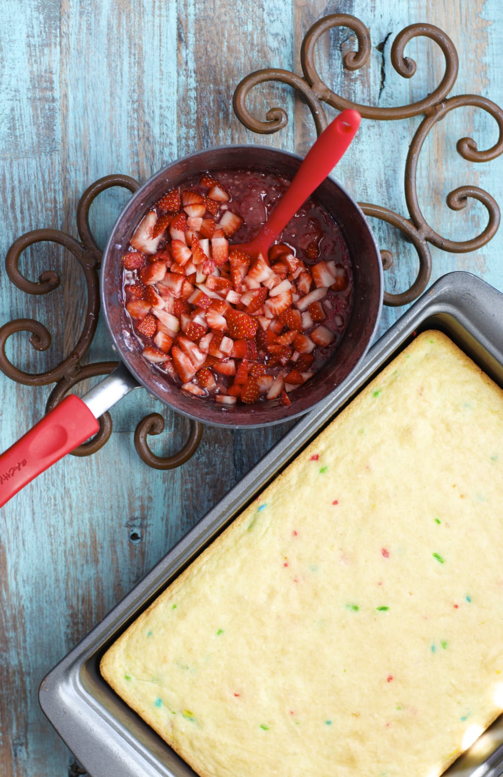 overhead view of gluten free funfetti cake in a baking dish and a saucepan of fresh strawberry sauce