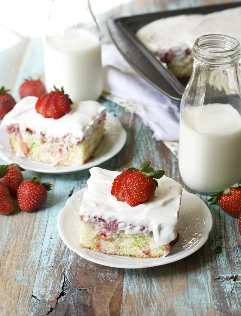 two slices of strawberry poke cake on white plates next to two glass jugs of milk. 