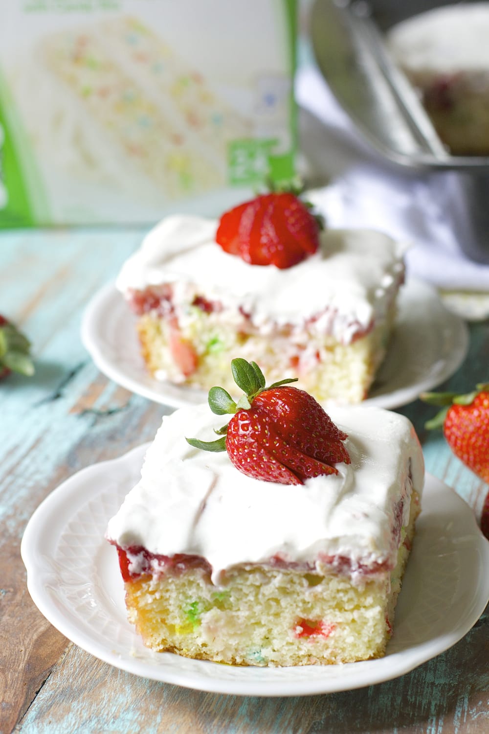 two slices of strawberry poke cake on white plates. a box of cake mix rests in the background. 