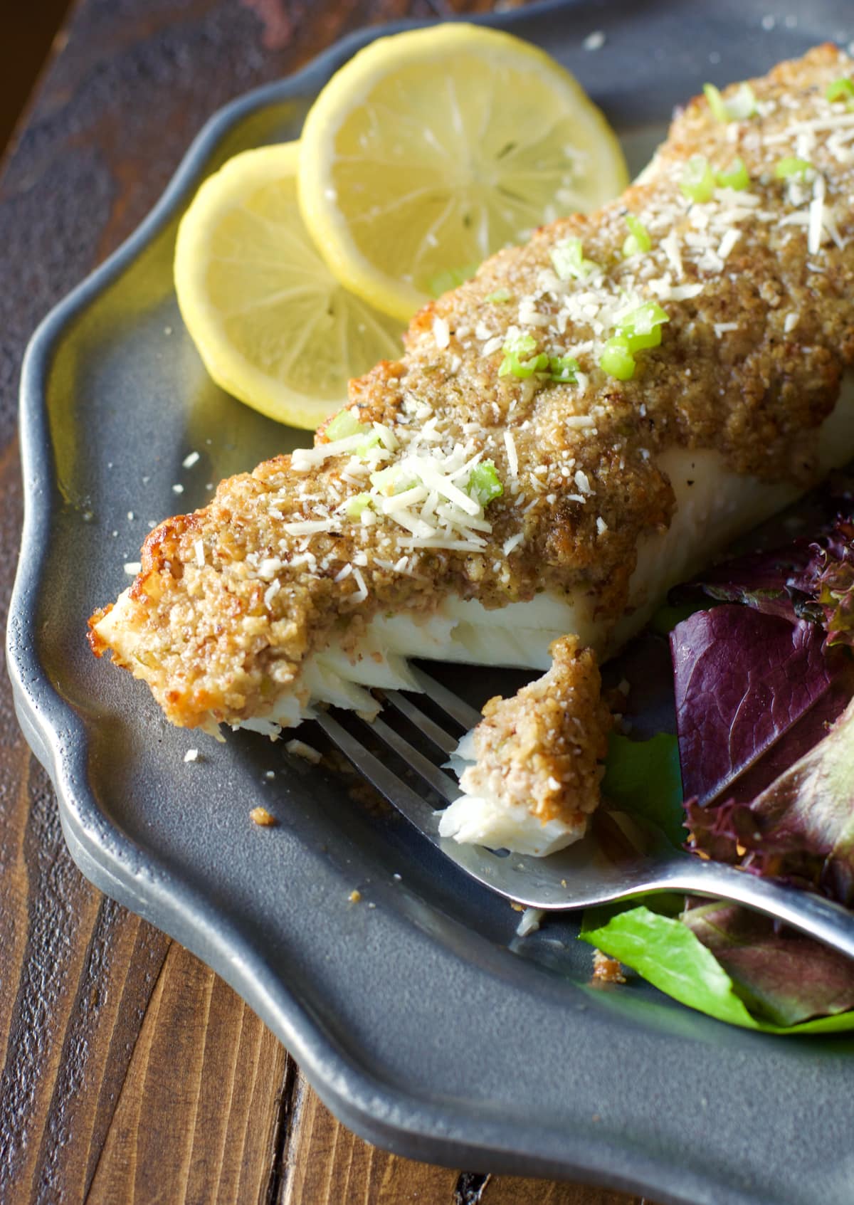 A pecan and parmesan crusted halibut fillet on a gray plate with lemon slices. 