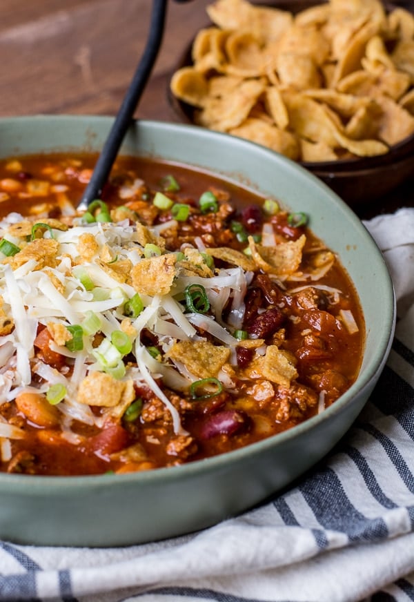 bowl of simple turkey chili topped with Fritos, cheese, and green onions. a bowl of fritos rests in the background 