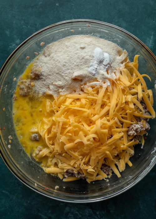 A mixing bowl filled with sausage meat, with a pile of shredded cheese, coconut flour, and baking powder on top, with some eggs