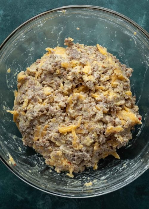 A glass mixing bowl filled with a mixture of sausage meat, cream cheese, cheddar cheese, eggs, coconut flour, and baking powder