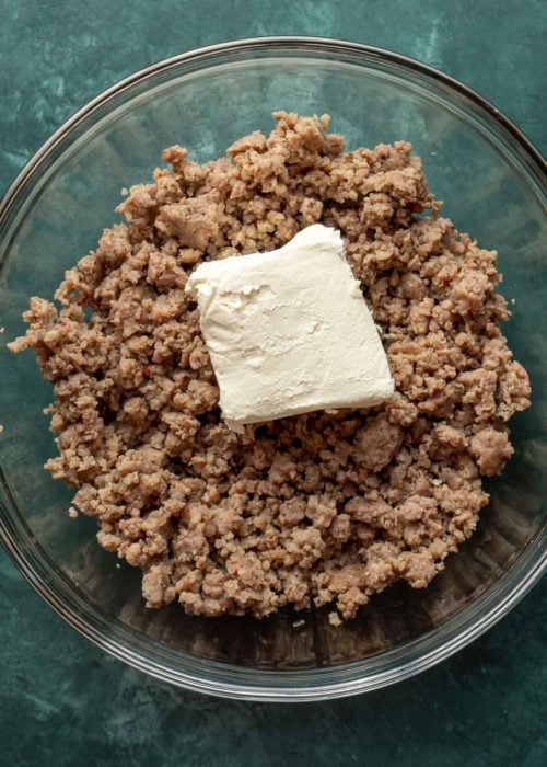 A mixing bowl with cooked sausage meat, with a block of cream cheese on top