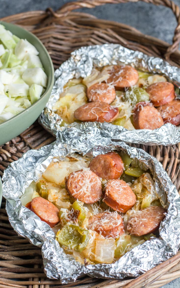Two cabbage with sausage foil packs on a wicker tray next to a small bowl of cabbage. 
