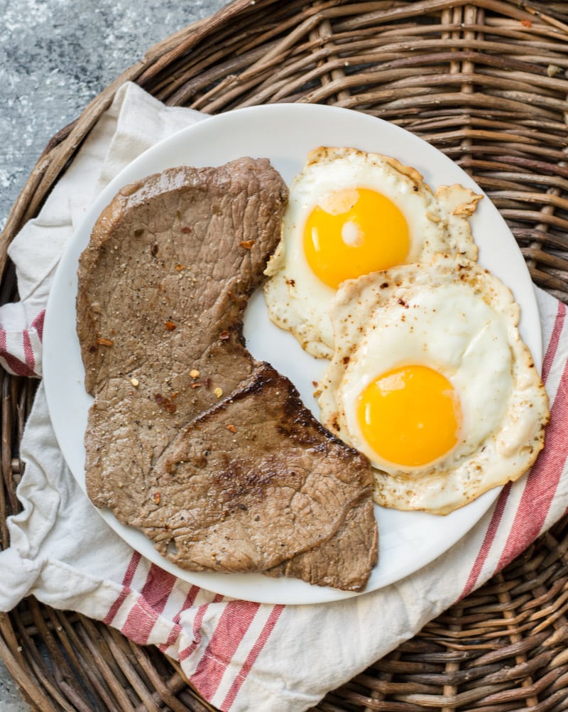 steak and eggs on a white plate, which is resting on a wicker tray 