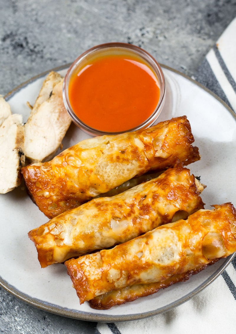 Three keto chicken and cheese taquitos on a white plate with a dish of buffalo sauce and some sliced chicken. 