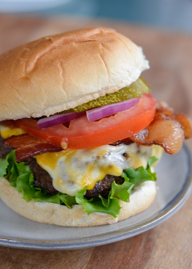 juicy air fryer cheeseburger with lettuce, bacon, tomato, red onion, and pickles on a bun on a white plate