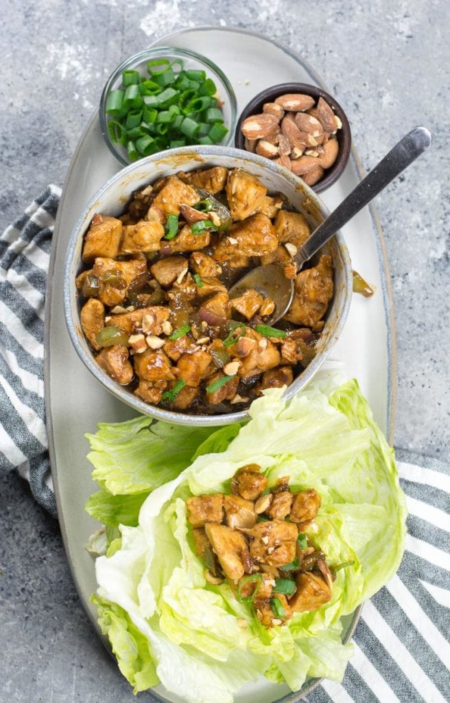 chicken salad in a bowl with a spoon on a platter with green onions, almonds, and a lettuce cup full of chicken salad