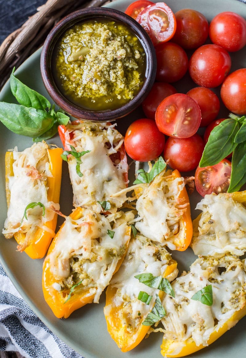 pesto chicken stuffed peppers on a green plate with tomatoes, basil, and pesto sauce in a small bowl