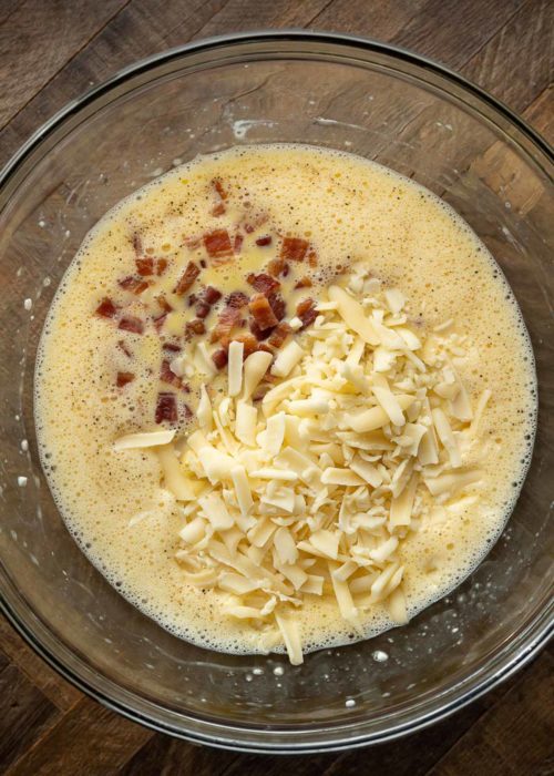 A mixing bowl with pieces of bacon and shredded cheese on top of a mixture of eggs and cottage cheese