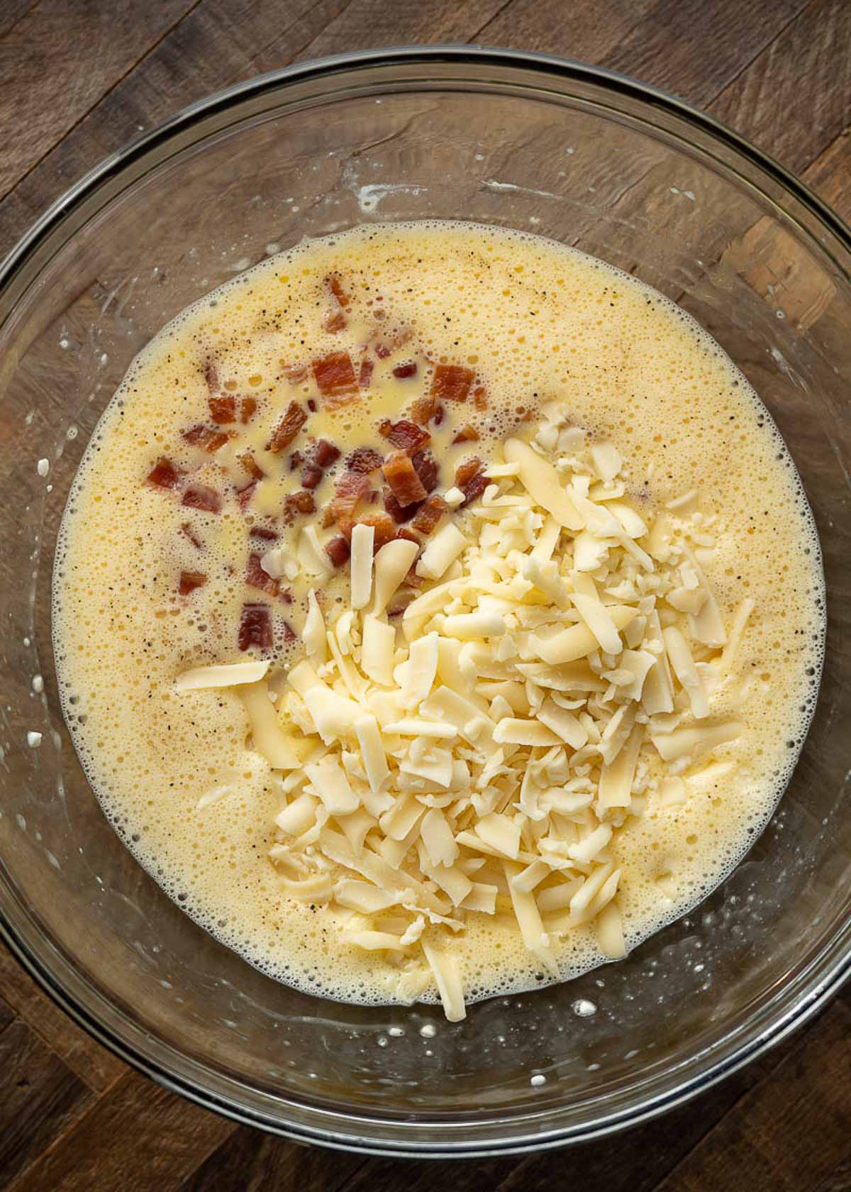 A mixing bowl with pieces of bacon and shredded cheese on top of a mixture of eggs and cottage cheese