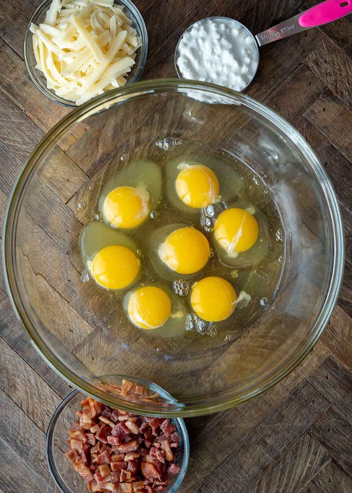 Overhead view of the ingredients needed for egg bites: a bowl of eggs, a bowl of bacon pieces, a bowl of shredded. gruyere cheese, and a measuring cup of cottage cheese