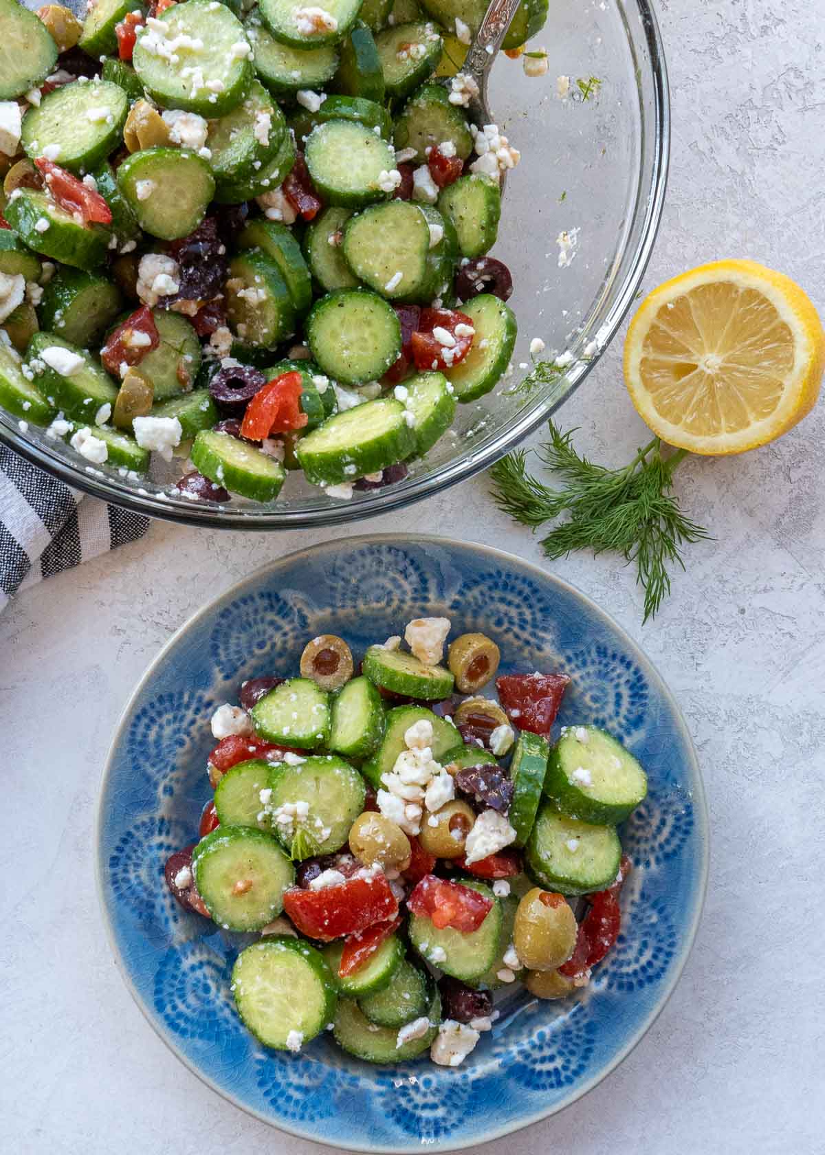 Overhead view of cucumber salad in glass bowl and on plate with sprig of dill and lemon half