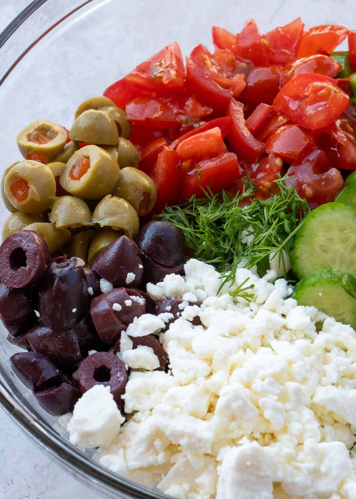 Glass mixing bowl with cucumbers, feta, dill, olives, and tomatoes