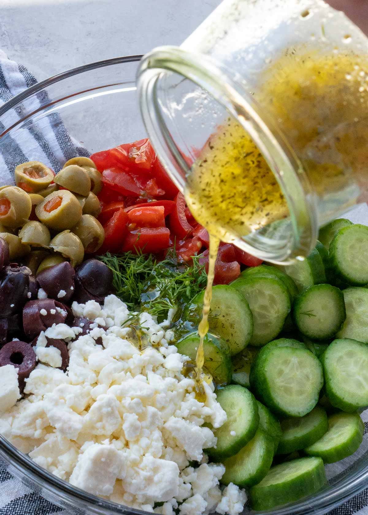 Pouring jar of salad dressing into bowl of cucumbers, feta, olives, and tomatoes