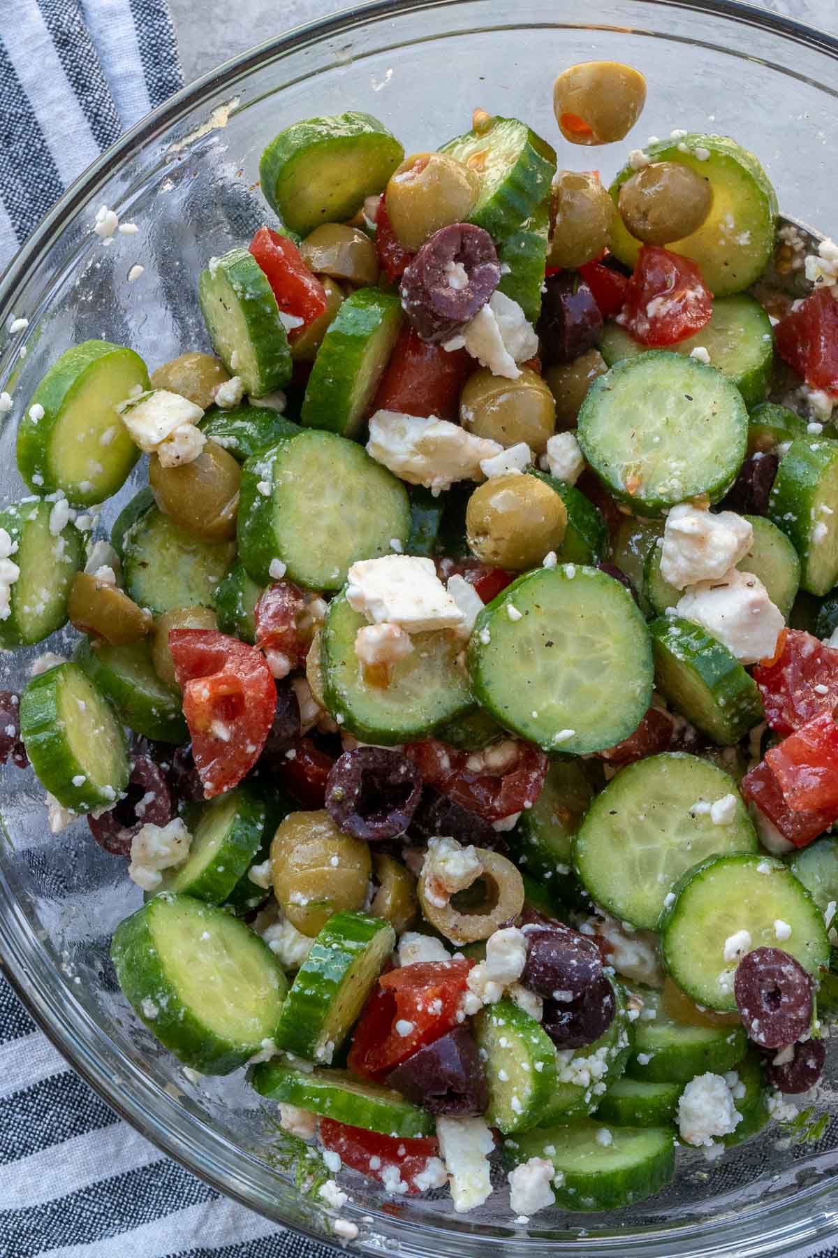 Overhead view of cucumber salad with olives, tomatoes, and feta in glass bowl