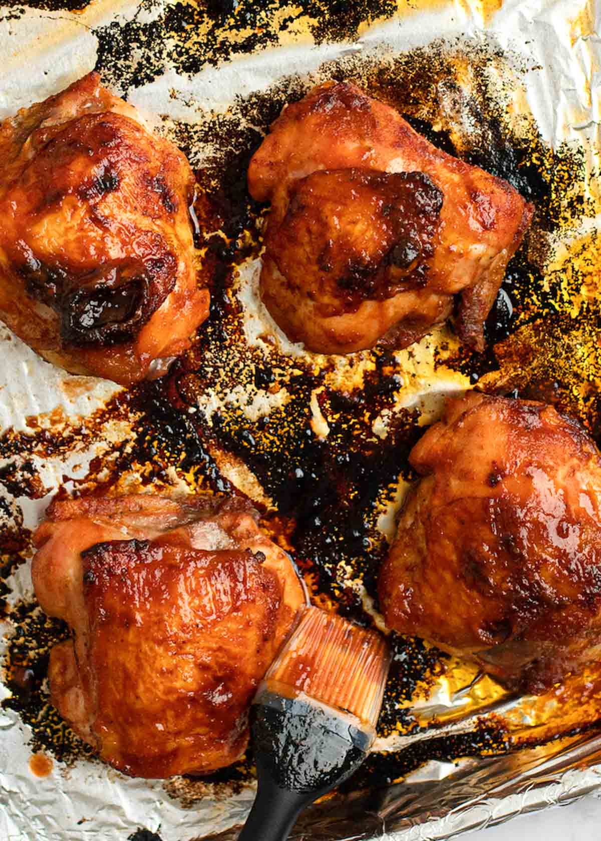 homemade barbecue sauce being brushed onto baked chicken thighs on a foil-lined baking pan