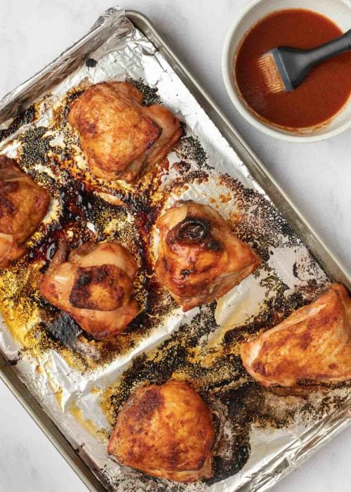 baked chicken thighs on an aluminum foil-lined baking pan beside a small bowl of marinade with a brush
