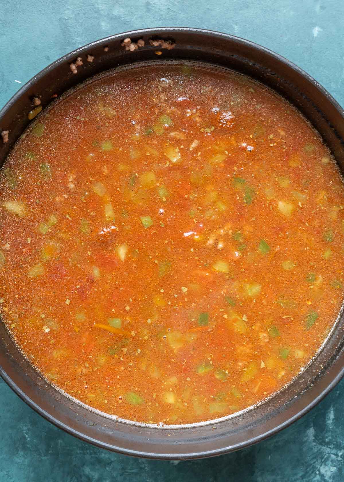 dutch oven two-thirds filled with beef broth, ground beef, tomatoes, tomato paste, green chilies, peppers, and seasonings