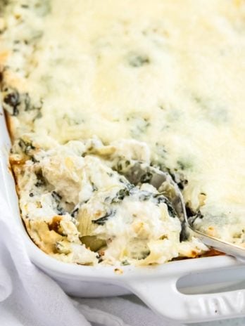 closeup of spinach artichoke dip in a white baking dish, being scooped out with a metal serving spoon