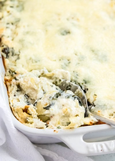 closeup of spinach artichoke dip in a white baking dish, being scooped out with a metal serving spoon