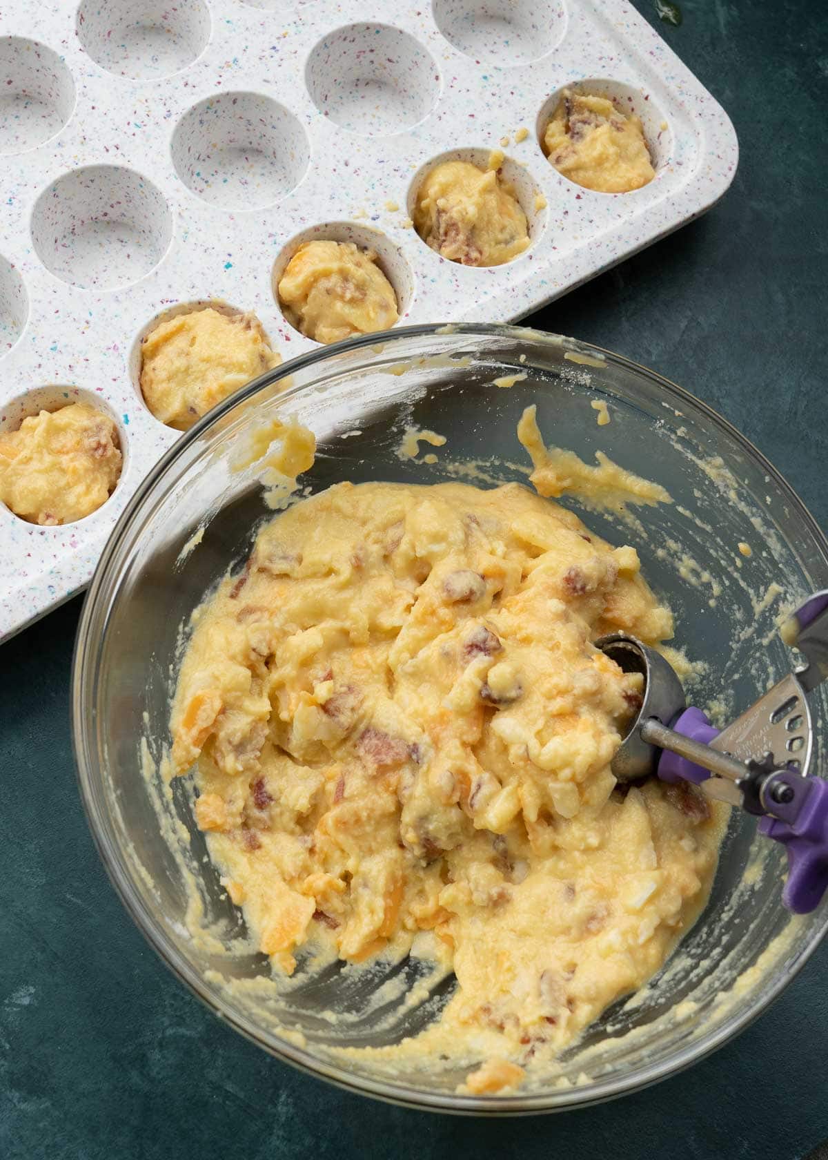 bowl of egg bite mixture with a scoop beside a mini muffin pan with 1/3rd of cavities filled.